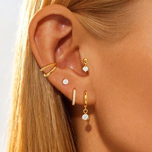 Puede incluir: Una colección de pendientes dorados, que incluye un brazalete doble para la oreja, un pendiente de botón, un pendiente de aro pequeño y un pendiente colgante con una gema transparente. Los pendientes se muestran en una oreja.