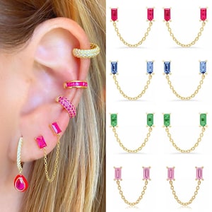 May include: A collection of gold-tone double-post earrings connected by a delicate chain, featuring rectangular baguette-cut stones in red, blue, green, and pink. The set includes a single hoop earring with a teardrop-shaped red stone.