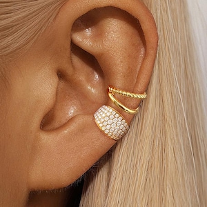 May include: Close-up of an ear adorned with three gold-colored ear cuffs. One cuff is a wide band covered in small, clear stones. The other two are simple, curved bands, one with a twisted rope design.