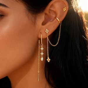 May include: A selection of gold-coloured earrings and ear cuffs. The earrings showcase diverse designs, including a chain connecting two floral-shaped studs, a hoop with dangling geometric shapes, and a stud with a small flower design.