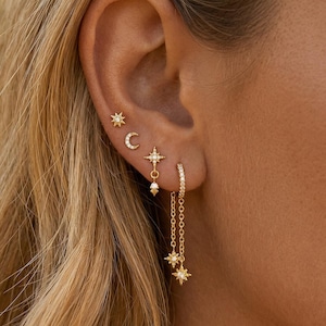 May include: A selection of gold earrings, featuring star and crescent moon studs, a dangling star earring with a small pearl, and a hoop earring with a chain and star charms. The earrings are embellished with tiny, sparkling stones.