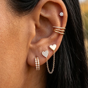 May include: A gold ear stack featuring a single crystal stud, a triple-band ear cuff with crystals, a double-row crystal hoop, and a heart-shaped stud connected by a delicate gold chain to a solid gold heart stud, all worn on a single ear.
