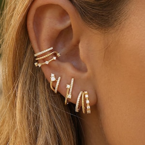 May include: A collection of gold-coloured ear jewellery, including multiple earrings and ear cuffs. The set features a triple-band ear cuff, a curved earring with clear stones, a straight bar earring, and a double-band hoop earring, all with small, clear stones.