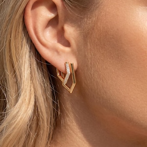 Può includere: Orecchini geometrici color oro con un accento scintillante simile a un diamante. Gli orecchini hanno un design angolare unico, con una combinazione di linee rette e spazi aperti. Gli orecchini sono mostrati su un orecchio, evidenziando la loro forma e il loro stile.