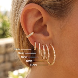 May include: Gold-toned hoop earrings and ear cuff, encrusted with small, clear stones. Sizes range from 13mm to 26mm. The earrings are displayed on an ear, showcasing the different sizes and styles available.