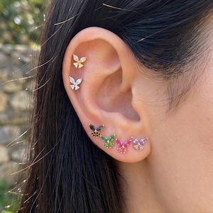 Puede incluir: Una colección de seis pendientes de botón con forma de mariposa dispuestos a lo largo del cartílago y el lóbulo de la oreja. Los pendientes presentan piedras de varios colores, incluyendo transparentes, negras, verdes, rosas y moradas.