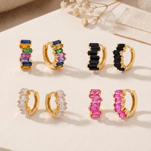 May include: Four pairs of gold hoop earrings featuring baguette-cut gemstones. One pair has multicoloured stones, one has black stones, one has clear white stones, and one has bright pink stones. Each hoop is secured with a hinged clasp mechanism.