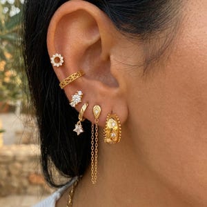 Peut inclure: Plusieurs boucles d'oreilles dorées, dont une pendante en forme d'étoile, une créole avec des étoiles et une petite puce d'oreille. Les boucles d'oreilles sont portées sur l'oreille, présentant divers motifs et textures.