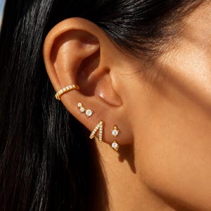 Puede incluir: Primer plano de una oreja adornada con múltiples piezas de joyería dorada. La oreja presenta un brazalete de oreja dorado, un grupo de tres pequeños tachuelas de diamantes, un pendiente en forma de V, un solo tachuela y un pequeño pendiente de aro con un diamante colgante.