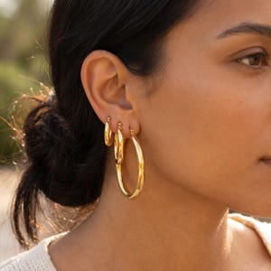 May include: A close-up of a person's ear adorned with three gold hoop earrings of varying sizes. The earrings have a polished, reflective surface, catching the light. The largest hoop is approximately 2 inches in diameter.