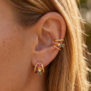 May include: Gold-toned ear cuff and earring set. The ear cuff wraps around the ear with two bands, adorned with colorful gemstones. The matching earring has a curved design, also featuring a row of colorful gemstones.