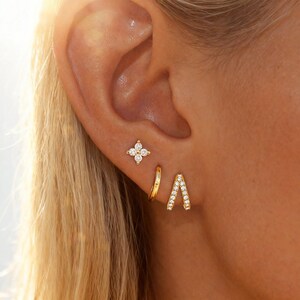 May include: A close-up of an ear adorned with three gold earrings. One is a small flower shape with clear stones, another is a simple hoop, and the third is a gold V-shaped earring with clear stones. The ear is set against a background of blonde hair.