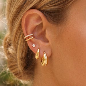 May include: A collection of gold earrings and ear cuffs. The ear cuffs feature a double band design with small, clear stones. A small, round stud earring with a single clear stone is also visible, along with a pair of teardrop-shaped earrings and a pair of hoop earrings with clear stones.