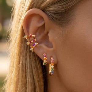 May include: A set of gold-toned earrings and ear cuffs featuring small, round, multi-colored gemstones. The collection includes a long rectangular hoop, a small circular hoop, and three stacked ear cuffs, all adorned with vibrant rainbow-colored stones.