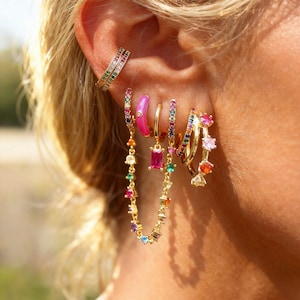 Peut inclure: Ensemble de boucles d'oreilles dorées ornées de pierres précieuses colorées. L'ensemble comprend des créoles, des clous et une chaîne, avec des pierres roses, violettes, vertes, oranges et transparentes. Un ear cuff arc-en-ciel est également visible.