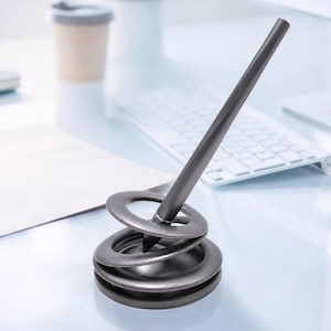 Könnte beinhalten: Ein dunkelgrauer Metallstift, der schräg in einem spiralförmigen Stiftehalter ruht. Der Halter besteht aus drei gestapelten, kreisförmigen Ringen, die nach oben hin kleiner werden und eine gewundene Struktur bilden, die den Stift aufrecht hält.