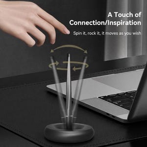 Könnte beinhalten: Ein schwarzes Schreibtischzubehör mit einem sich drehenden, silberfarbenen Zentralelement. Die Basis ist rund und dunkel. Der Text lautet "A Touch of Connection/Inspiration" und "Spin it, rock it, it moves as you wish."