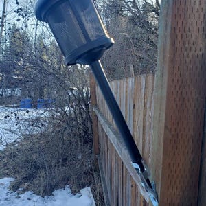 May include: A black outdoor solar-powered lamp attached to a wooden fence. The lamp has a cylindrical, mesh-covered light housing and a long, black pole. The fence is made of weathered wood, and snow covers the ground.
