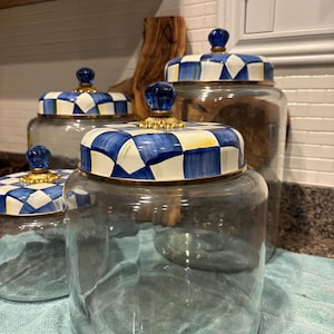 May include: A set of four glass jars with decorative lids. The lids feature a blue and white checkered pattern with a gold rim and a blue glass knob. The jars are empty, and the set is displayed on a blue towel.