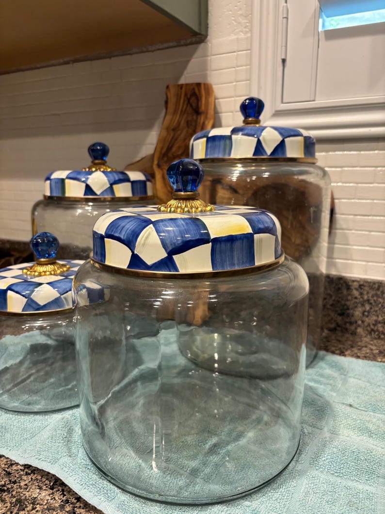 May include: A set of four glass jars with decorative lids. The lids feature a blue and white checkered pattern with a gold rim and a blue glass knob. The jars are empty, and the set is displayed on a blue towel.
