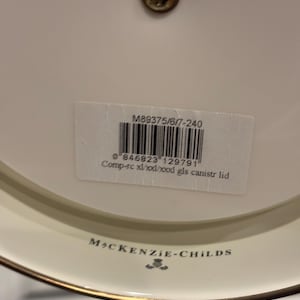 May include: A close-up of a white ceramic lid with a gold rim and a small brass screw. The lid has a label with a barcode and the text "M89375/6/7-240" and "MACKENZIE-CHILDS".