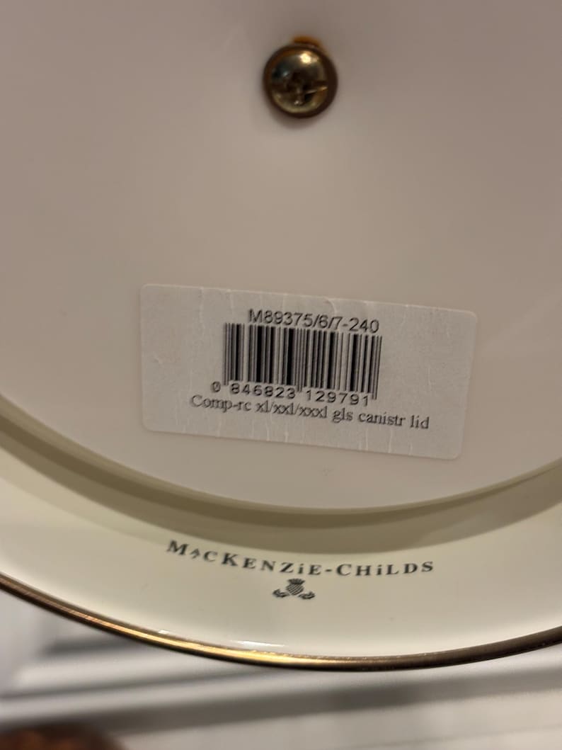 May include: A close-up of a white ceramic lid with a gold rim and a small brass screw. The lid has a label with a barcode and the text "M89375/6/7-240" and "MACKENZIE-CHILDS".