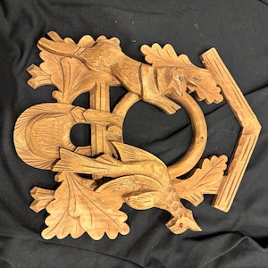 May include: A carved wooden wall hanging featuring a squirrel and a bird. The squirrel is leaping, and the bird is perched, both amidst oak leaves. The piece has a natural wood finish and includes a ring and a geometric element.