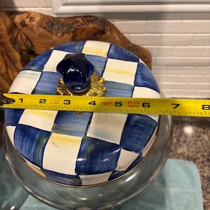 May include: A glass jar with a lid featuring a blue and white checkered pattern. The lid has a dark blue knob and measures approximately 7 inches in diameter. The jar is clear and has a gold-colored rim.