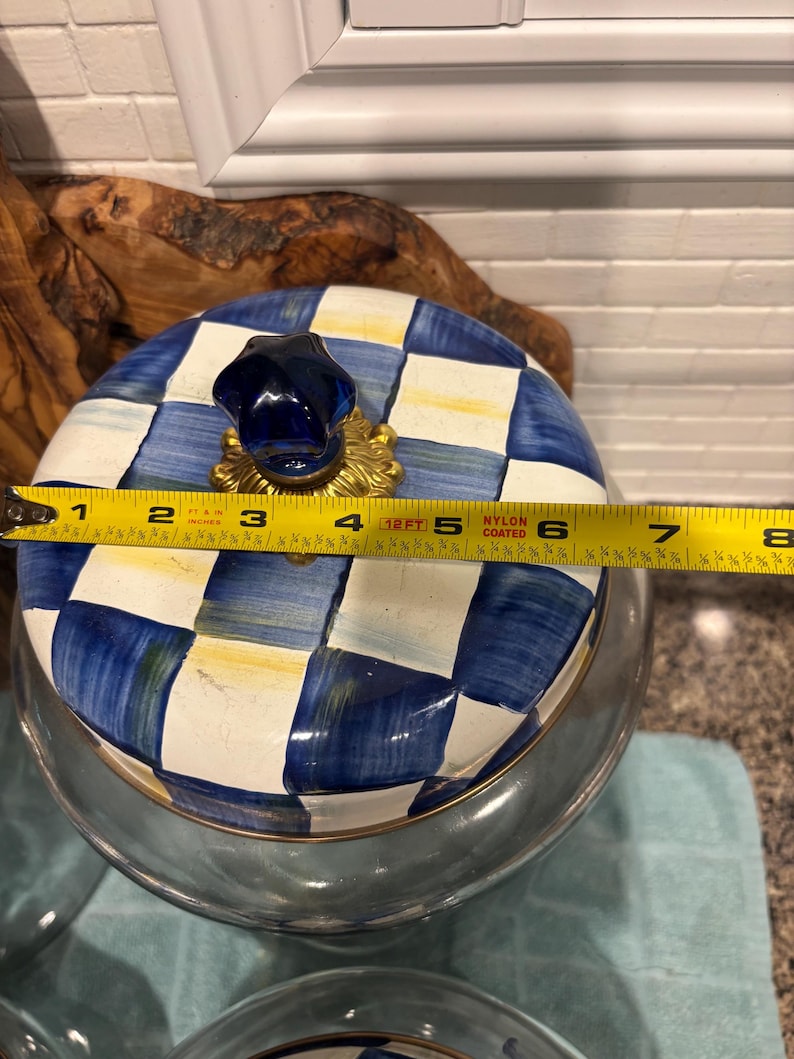 May include: A glass jar with a lid featuring a blue and white checkered pattern. The lid has a dark blue knob and measures approximately 7 inches in diameter. The jar is clear and has a gold-colored rim.