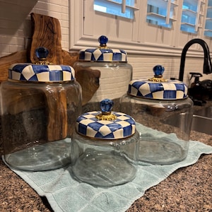 May include: Four clear glass jars with blue and white checkered lids and gold accents. The jars are different sizes and sit on a light blue towel. Each jar has a blue glass knob on top.