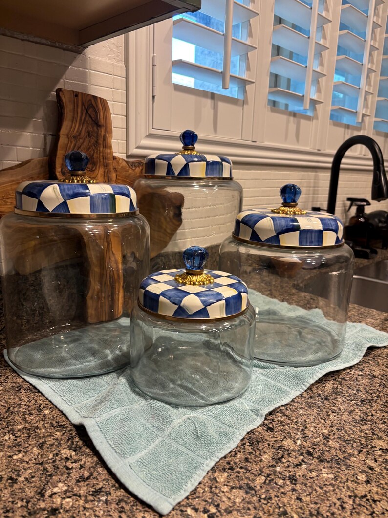 May include: Four clear glass jars with blue and white checkered lids and gold accents. The jars are different sizes and sit on a light blue towel. Each jar has a blue glass knob on top.