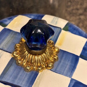 May include: A blue and white checkered ceramic lid with a gold-colored decorative base and a dark blue glass knob. The knob is shaped like a star. The lid has a yellow accent color.