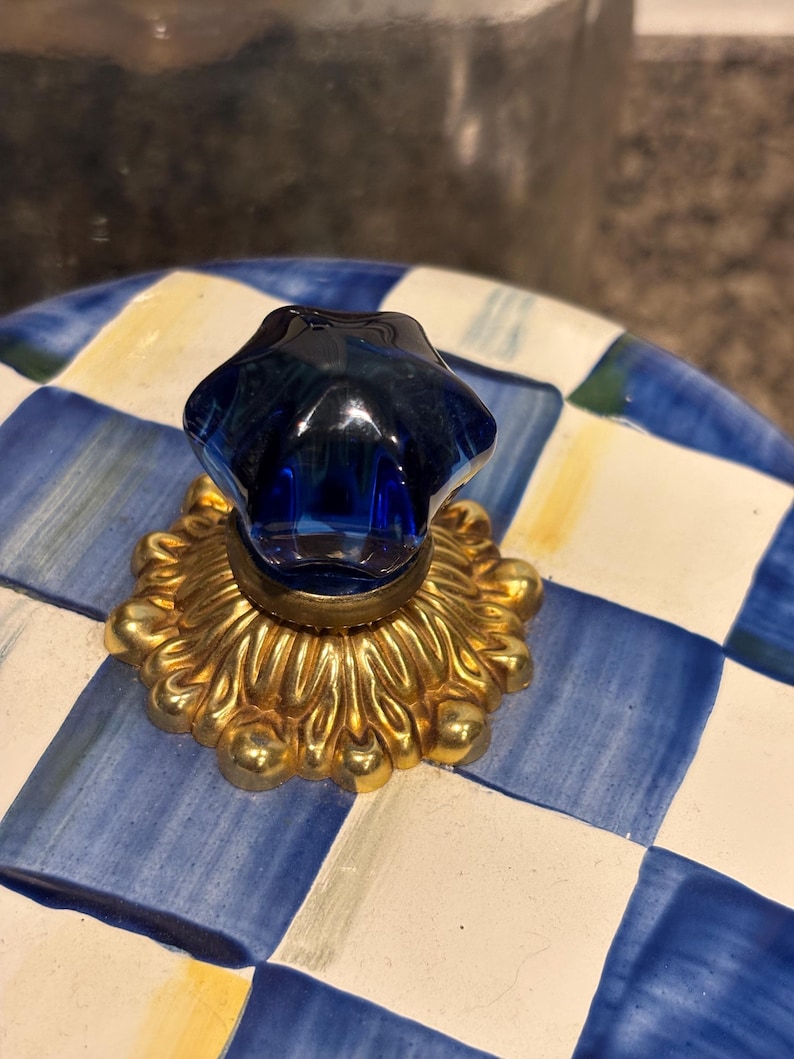 May include: A blue and white checkered ceramic lid with a gold-colored decorative base and a dark blue glass knob. The knob is shaped like a star. The lid has a yellow accent color.