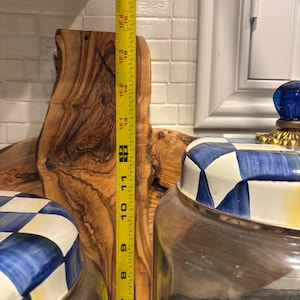 May include: A wooden cutting board with a natural grain pattern stands next to two glass jars with blue and white checkered lids. A yellow measuring tape shows the cutting board is approximately 16 inches tall.
