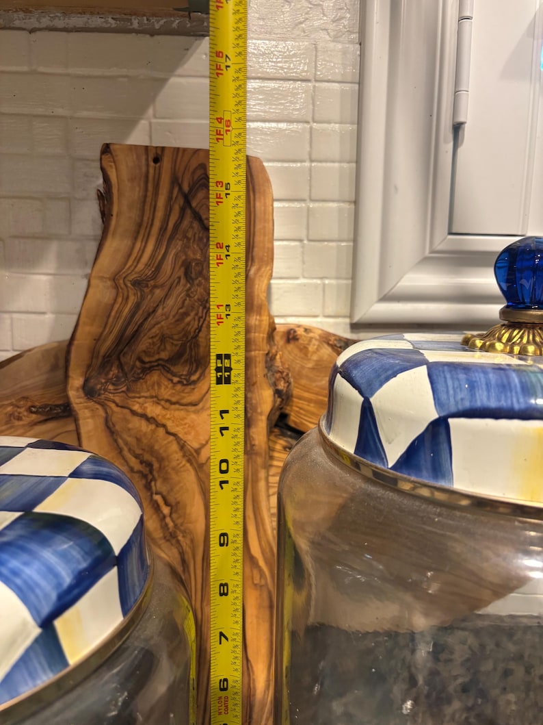 May include: A wooden cutting board with a natural grain pattern stands next to two glass jars with blue and white checkered lids. A yellow measuring tape shows the cutting board is approximately 16 inches tall.