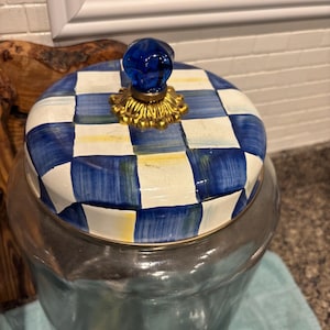 May include: A glass jar with a lid featuring a blue and white checkered pattern. The lid has a gold-colored base and a blue glass knob. The jar is likely used for storage and has a silver-colored rim.