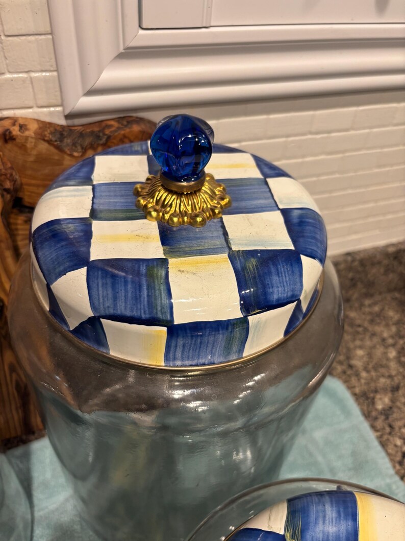 May include: A glass jar with a lid featuring a blue and white checkered pattern. The lid has a gold-colored base and a blue glass knob. The jar is likely used for storage and has a silver-colored rim.