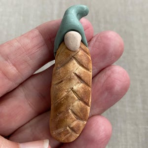 May include: A handmade clay gnome figurine. The gnome has a light brown body with a textured pattern, a light green hat, and a beige face. The figurine is held in a hand.