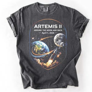 May include: Dark gray t-shirt featuring a graphic of the Artemis II mission, with Earth, the Moon, and a rocket. The text reads "ARTEMIS II AROUND THE MOON AND BACK - April 1, 2026". The shirt has a crew neck and short sleeves.