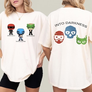 Könnte beinhalten: Cremefarbene T-Shirts mit grafischen Designs. Die Vorderseite des Shirts zeigt drei Cartoonfiguren mit den Texten: "JUNIOR TROCCI", "THE FACELESS MAN" und "MR.STRICKLAND". Auf der Rückseite des Shirts steht der Text "INTO DARKNESS" und drei maskenartige Figuren.