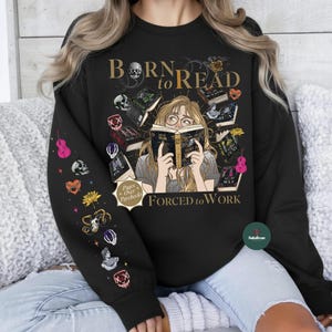 May include: Black sweatshirt with the text "Born to Read Forced to Work" and a graphic of a person reading surrounded by books. The left sleeve has various colorful illustrations. The design includes a skull and other decorative elements.