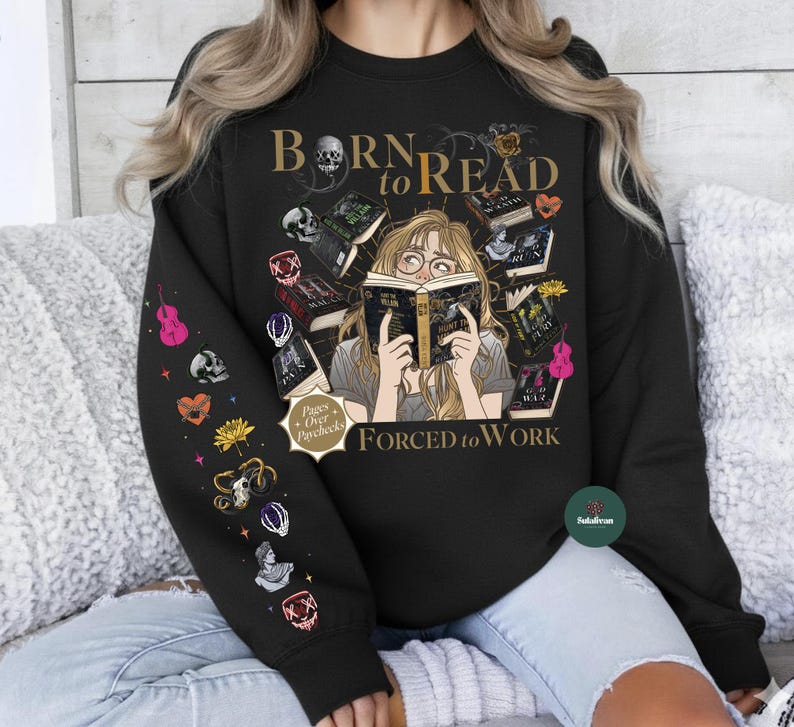 May include: Black sweatshirt with the text "Born to Read Forced to Work" and a graphic of a person reading surrounded by books. The left sleeve has various colorful illustrations. The design includes a skull and other decorative elements.