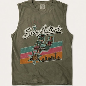 Puede incluir: Una camiseta sin mangas de color verde oliva con el texto San Antonio en letra blanca. Un logotipo de los Spurs relleno de patrones coloridos se sit&uacute;a sobre franjas horizontales en verde azulado, rosa y naranja, con una silueta urbana.