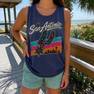 Puede incluir: Una camiseta sin mangas de color azul marino con un estampado gr&aacute;fico que dice San Antonio, un logotipo estilizado de los Spurs, un bal&oacute;n de baloncesto, la silueta del horizonte de la ciudad y rayas horizontales en turquesa, rosa y naranja.
