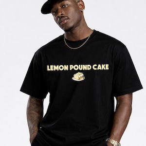 Puede incluir: Camiseta negra con la frase "LEMON POUND CAKE" en letras doradas. Debajo, una pequeña ilustración de una porción de bizcocho de limón. La camiseta es de cuello redondo.