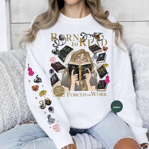 May include: White sweatshirt with the text "Born to Read, Forced to Work" and an illustration of a person reading surrounded by books. The left sleeve has various colorful graphics including a violin, skull, and flowers.