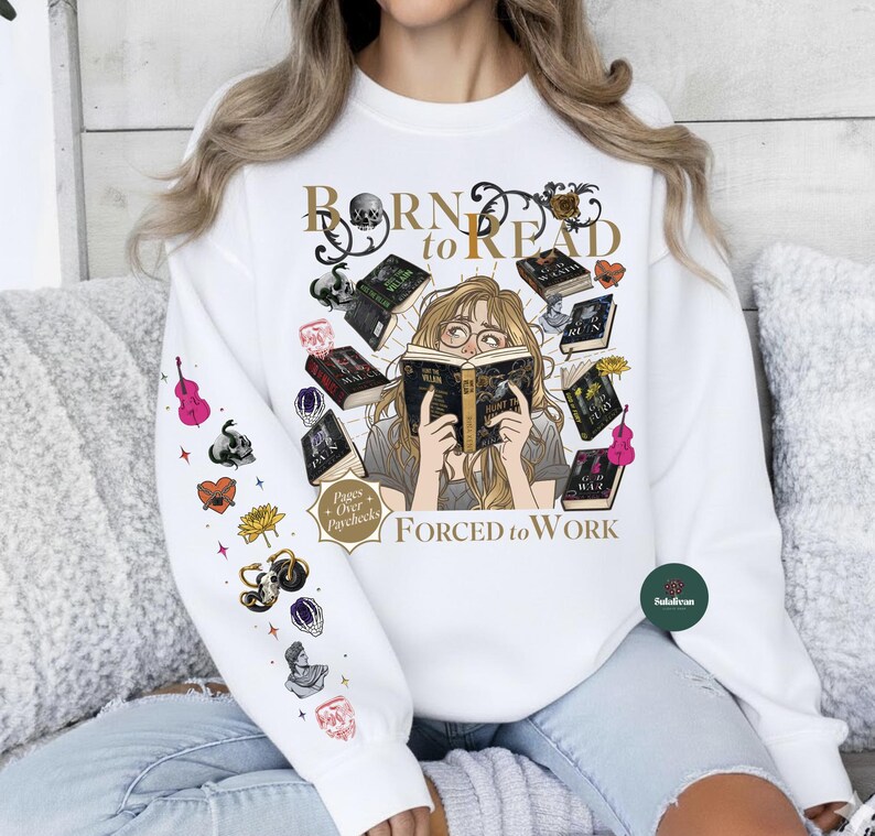 May include: White sweatshirt with the text "Born to Read, Forced to Work" and an illustration of a person reading surrounded by books. The left sleeve has various colorful graphics including a violin, skull, and flowers.