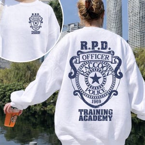 Może przedstawiać: Biała bluza z granatowym nadrukiem R.P.D. Officer Raccoon Police Training Academy z tyłu i z przodu. Projekt zawiera gwiazdę, rok 1969 i emblemat przypominający odznakę.