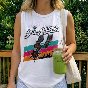 Puede incluir: Una camiseta sin mangas blanca con un estampado gr&aacute;fico que dice San Antonio en letra cursiva, un logotipo de los Spurs estilizado, una silueta del horizonte de la ciudad y rayas horizontales en turquesa, rosa y naranja, junto a un bolso de tela beige.