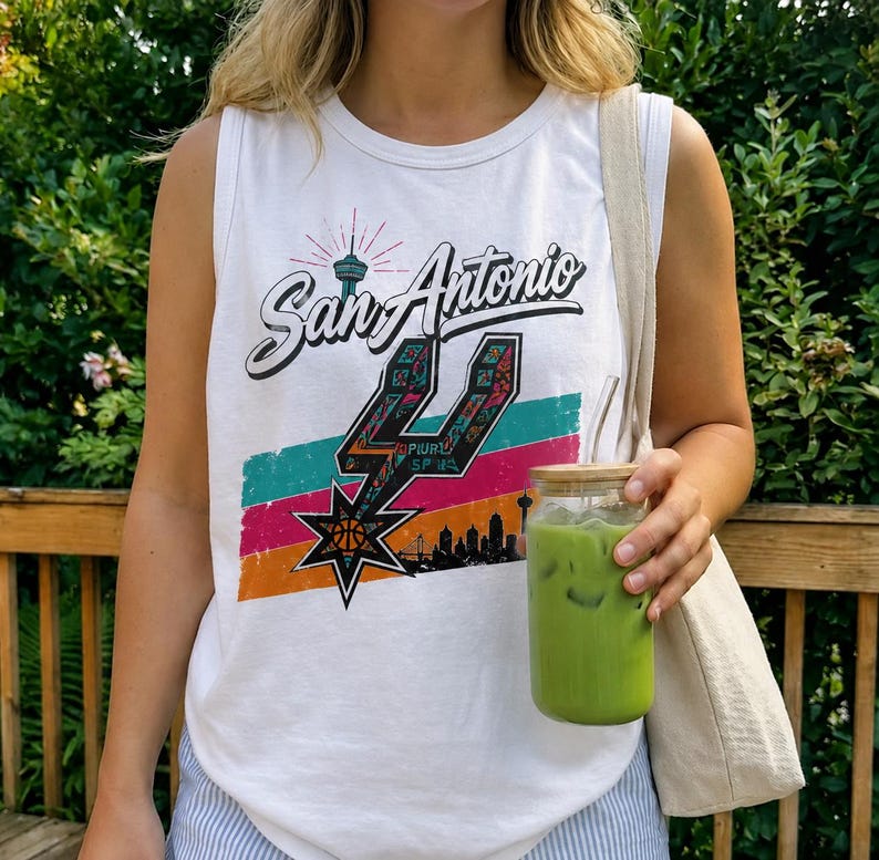 Puede incluir: Una camiseta sin mangas blanca con un estampado gr&aacute;fico que dice San Antonio en letra cursiva, un logotipo de los Spurs estilizado, una silueta del horizonte de la ciudad y rayas horizontales en turquesa, rosa y naranja, junto a un bolso de tela beige.